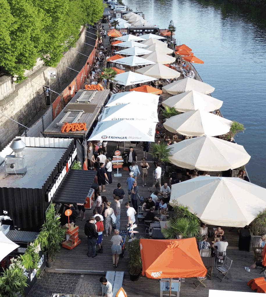 Eine beeindruckende Luftaufnahme oder erhöhte Perspektive des Würzburger Stadtstrands, die das belebte Areal mit Sonnenschirmen, Bars und vielen Gästen am Mainufer zeigt.