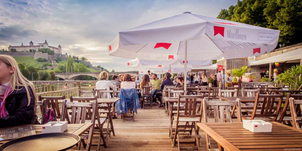 Gemütlicher Außenbereich mit Tischen und Schirmen am Würzburger Stadtstrand, wo Gäste Speisen und Getränke genießen – Gastronomie am Main.