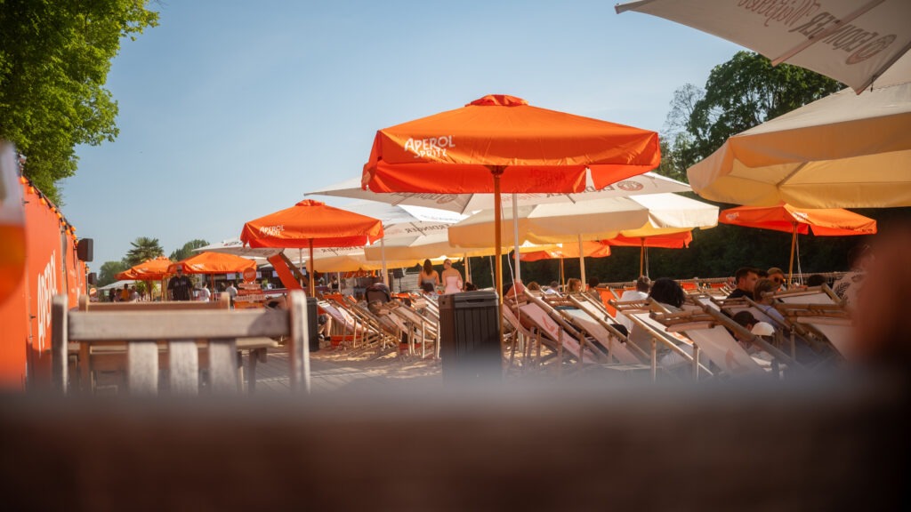 Strandbereich mit Aperol-Sonnenschirmen und Liegestühlen auf dem Stadtstrand Würzburg, bei strahlend blauem Himmel und warmem Wetter.