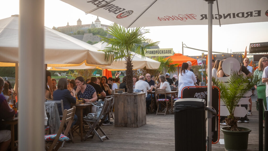 Menschen sitzen im Restaurantbereich des Stadtstrands Würzburg bei Sonnenuntergang, mit Blick auf die Festung Marienberg im Hintergrund