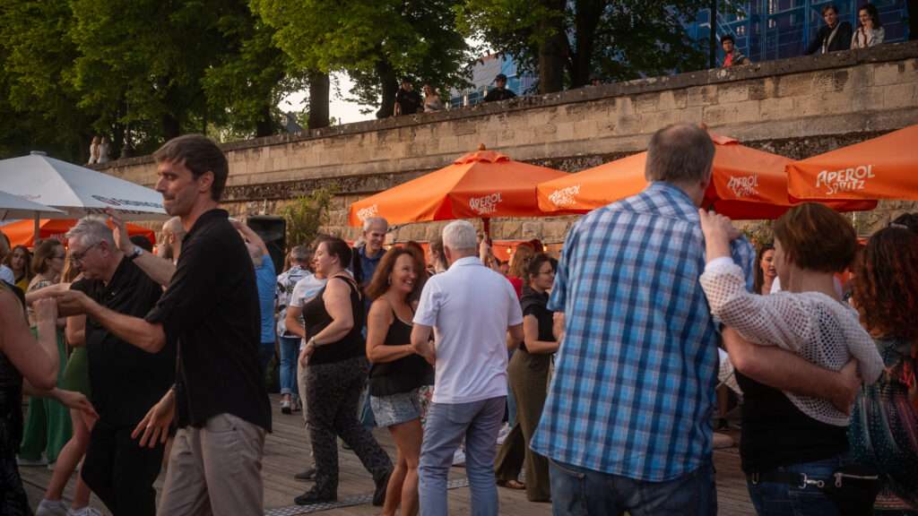 Menschen tanzen ausgelassen bei einem Event am Würzburger Stadtstrand, belebte Atmosphäre unter orangen Schirmen – Events in Würzburg