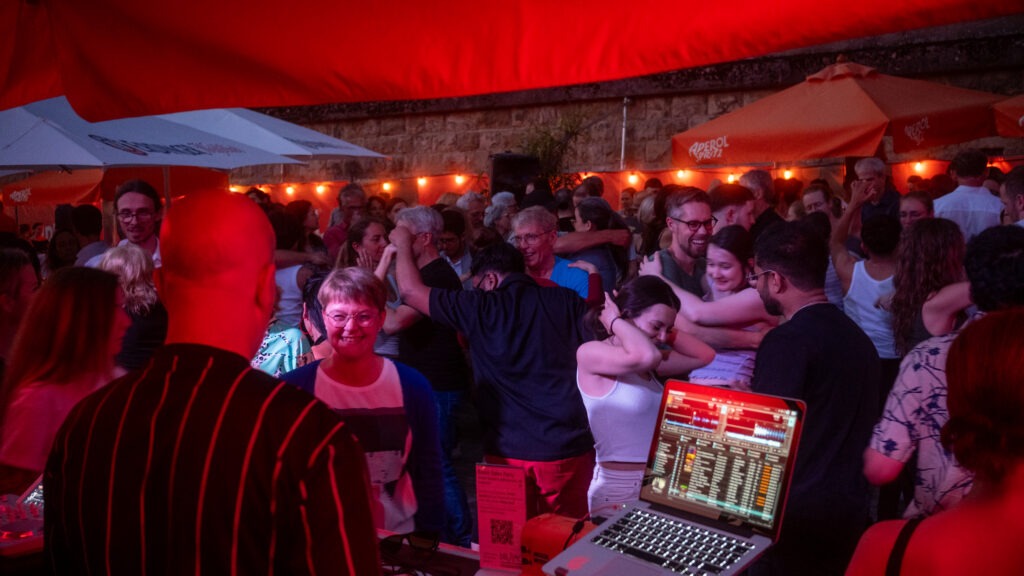 Menschen tanzen zu Live-DJ-Musik auf dem Stadtstrand Würzburg unter roten Sonnensegeln bei Abendstimmung